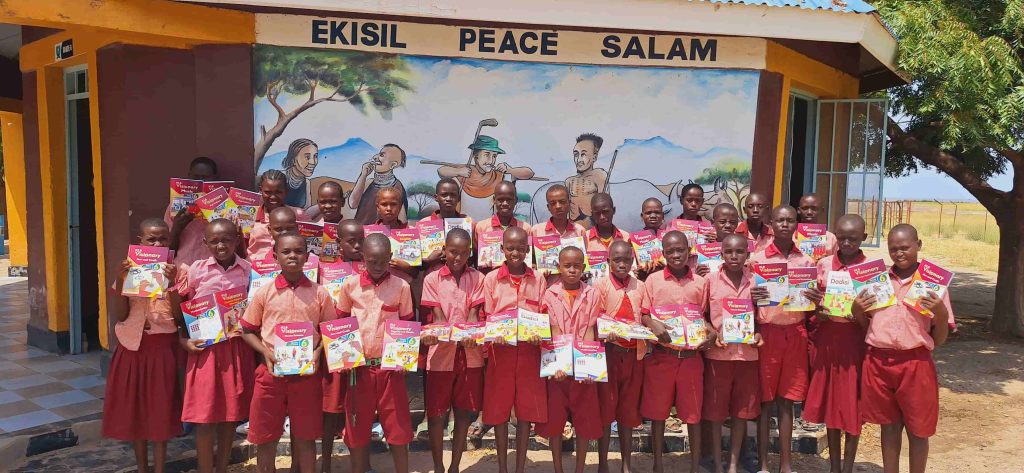 Group of smiling schoolchildren in red uniforms posing with colorful textbooks in front of a muraled wall at school.