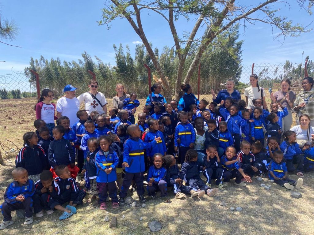 Group of children in blue uniforms posing for a photo outdoors with several adults behind them near a chain-link fence and a tree on a sunny day.