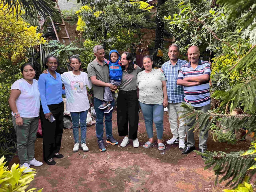 Group of nine people standing together in a garden, smiling among dense green plants in the background.