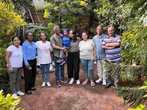 Group of nine people standing together in a garden, smiling among dense green plants in the background.