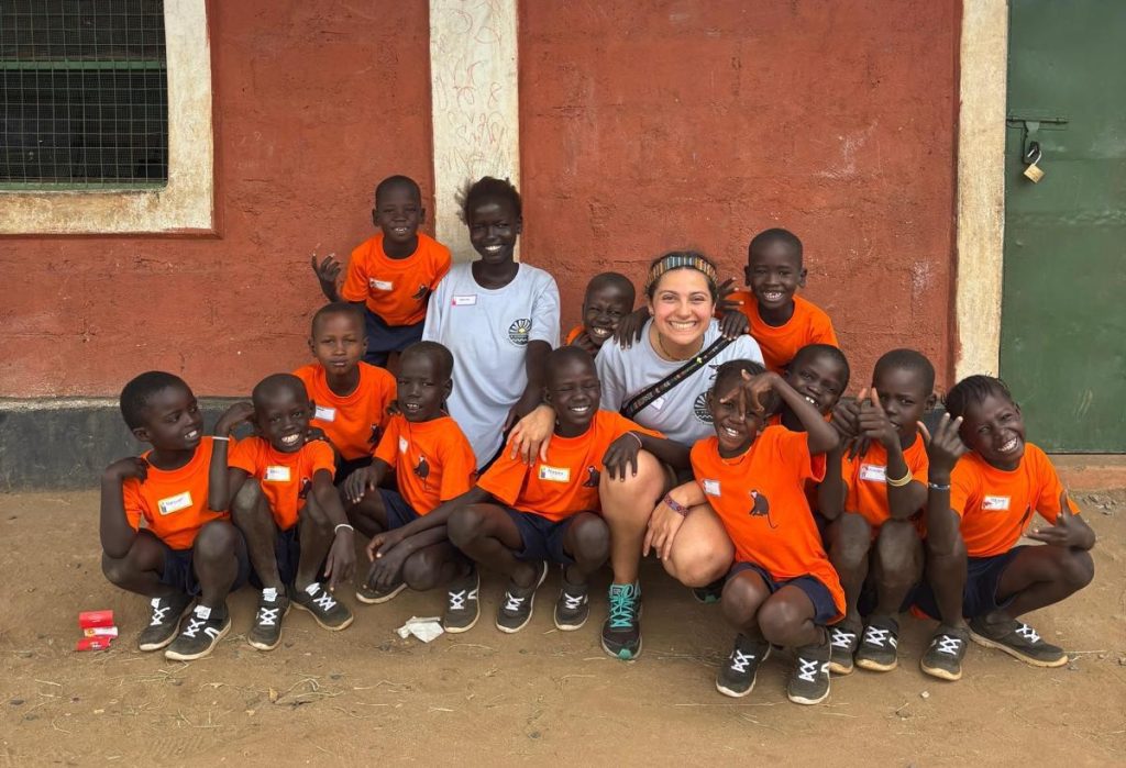 Group of smiling children in orange uniforms posing with a volunteer outside a red building.
