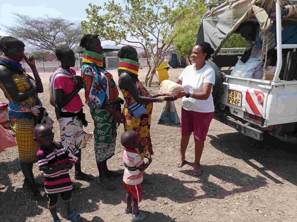 Woman in a white T-shirt hands a bag to a child as a group of villagers in colorful traditional clothing stand nearby near a white pickup truck in a dusty rural yard.