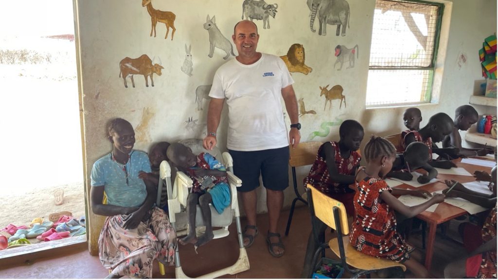 A volunteer stands in a classroom with children seated at a long table drawing; a child in a seat receives care from a supervising woman nearby as murals of animals decorate the wall behind them.