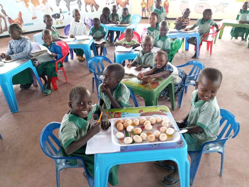 Group of smiling children in green uniforms sit at colorful plastic tables in a classroom, with a tray of eggs on the foreground table as they work on activities.