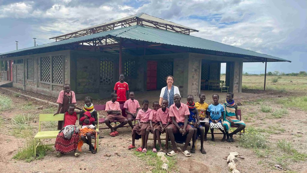 Group of children and a woman posing outside a single-story building with a blue metal roof in a rocky, rural area.