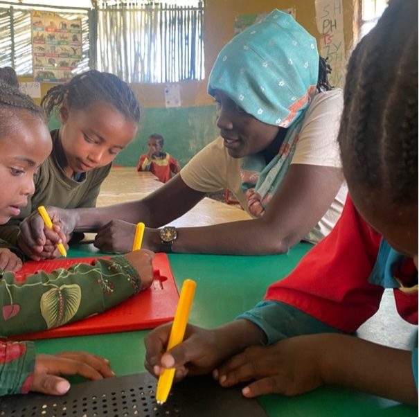 Teacher helps children draw with yellow markers at a green table in a bright classroom setting on a sunny day, outdoors-like space in the background.