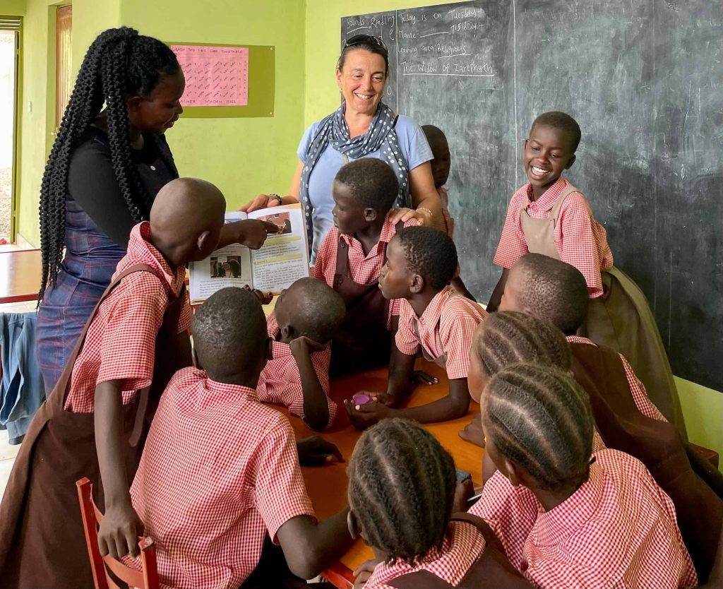 Teacher smiling and guiding students as they read a book together in a bright classroom; kids wear red checkered uniforms around a table.