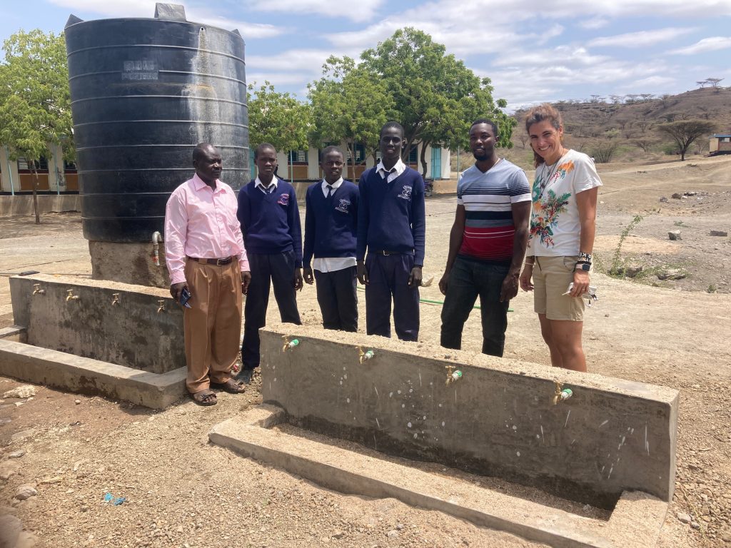 Six people stand beside a large black water tank in a dusty schoolyard, with a building and hills in the background.