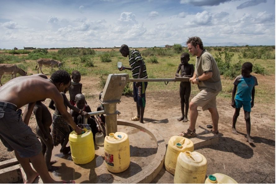 People gather around a manual hand-pump, filling yellow jerrycans with water in a dry, rural landscape.