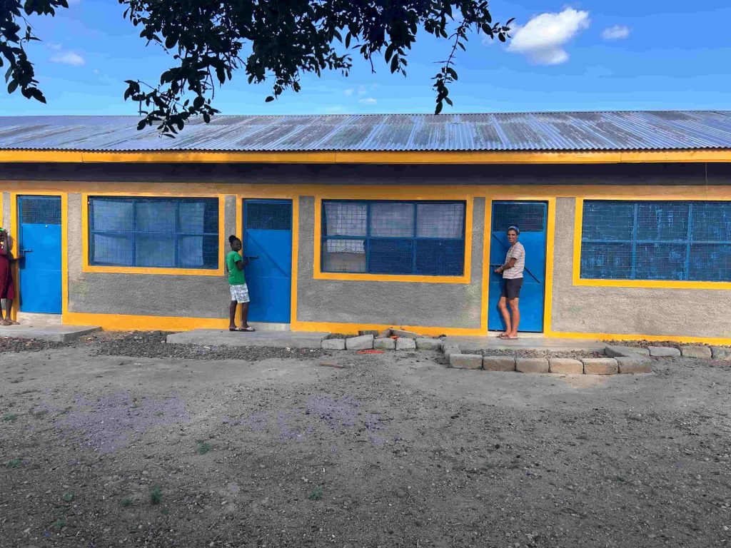 A single-story building with blue doors and windows framed by yellow trim; two people stand by the doors under a clear blue sky.