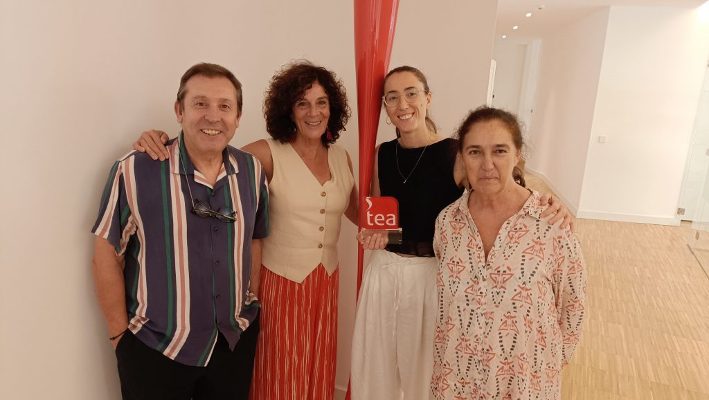 Four adults smiling at the camera indoors, standing beside a tall red sculpture and a small 'tea' award plaque.