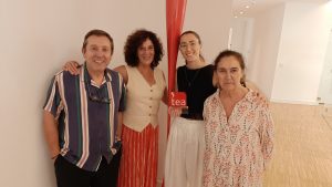 Four adults smiling at the camera indoors, standing beside a tall red sculpture and a small 'tea' award plaque.