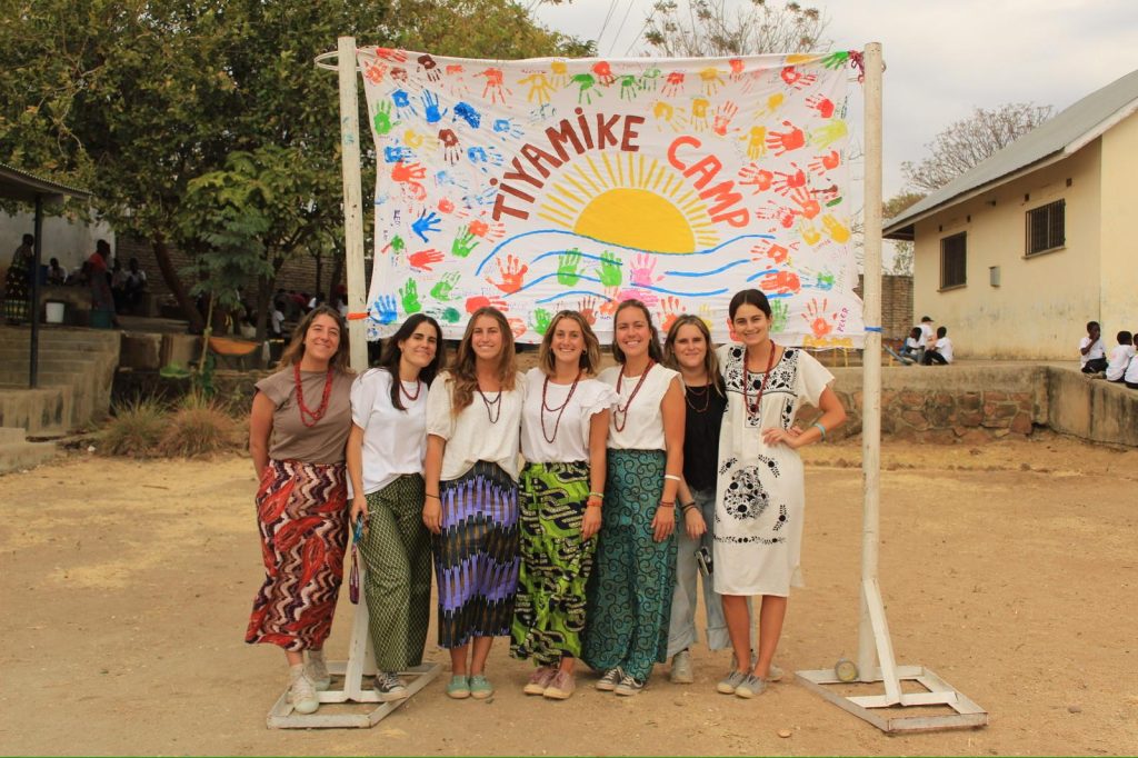 Seven young women stand in a row, wearing bright patterned skirts and white tops, posing in front of a colorful banner with a sun and handprints at an outdoor camp site.