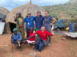 Group of men posing together outdoors in a rural village with thatched huts, smiling at the camera.