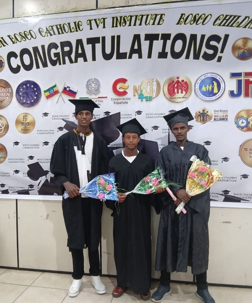 Three graduates in caps and gowns pose indoors in front of a large "CONGRATULATIONS!" banner, each holding a bouquet of flowers.