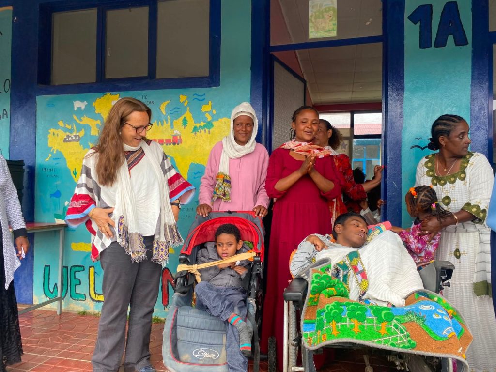 A diverse group of adults and two children in wheelchairs stand outside a colorful muraled building, smiling and engaging with each other.