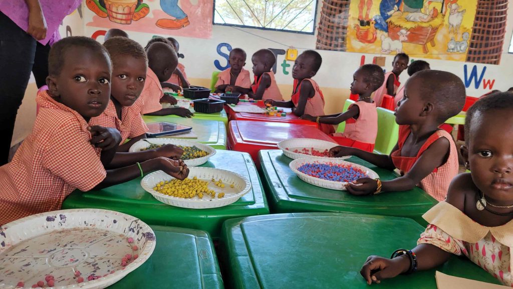 Group of young children at colorful tables sorting small beads into bowls during a classroom activity or craft session.