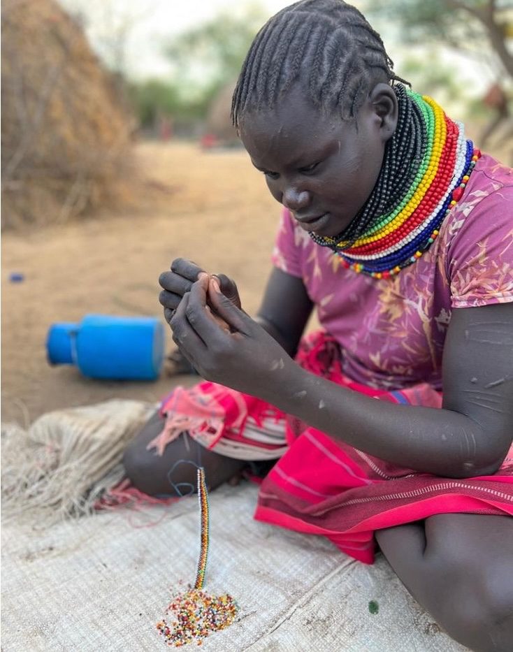 Maasai woman with braided hair and colorful bead necklaces threading beads outdoors on a mat.