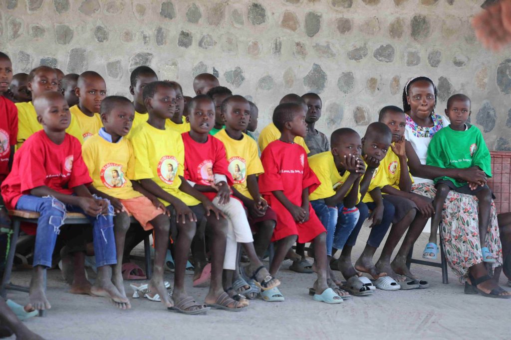A group of African children in yellow and red shirts seated on a bench, looking toward the left, with a woman in green on the right.