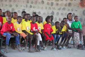 A group of African children in yellow and red shirts seated on a bench, looking toward the left, with a woman in green on the right.