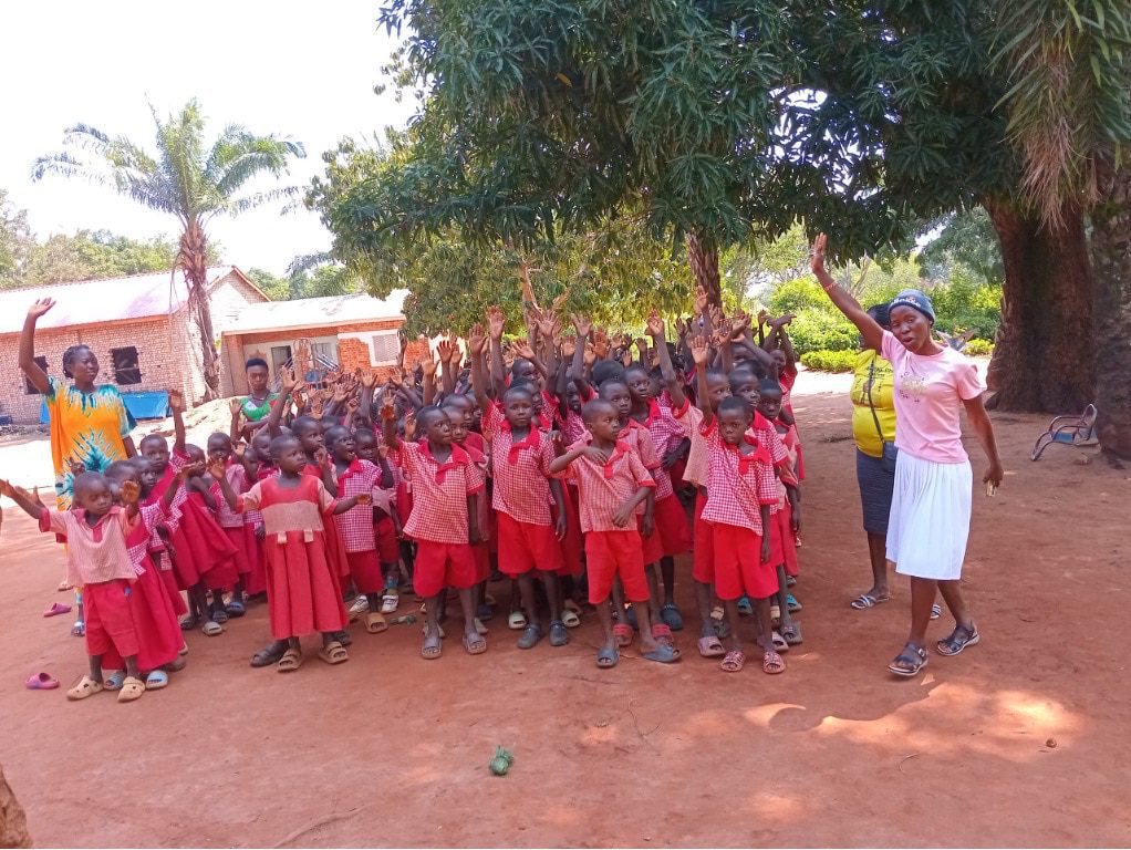 Group of young students in red uniforms waving hello outdoors under a large tree, with two adults on the right.