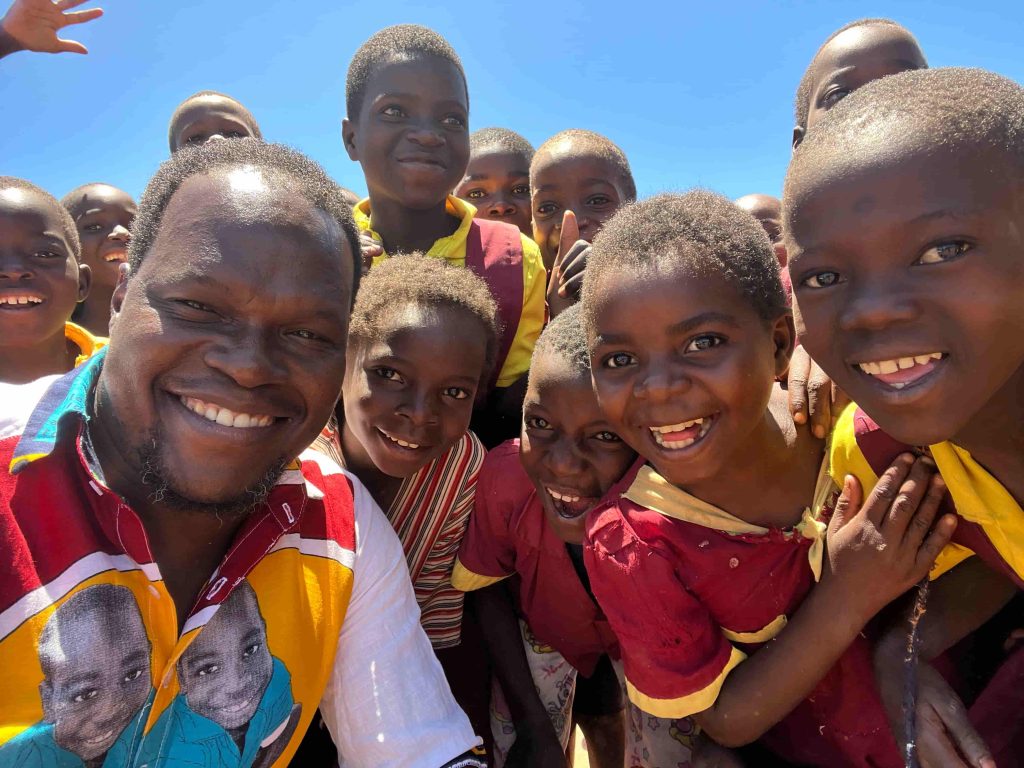 Group of smiling children taking a close-up selfie outside on a bright sunny day, wearing colorful shirts.