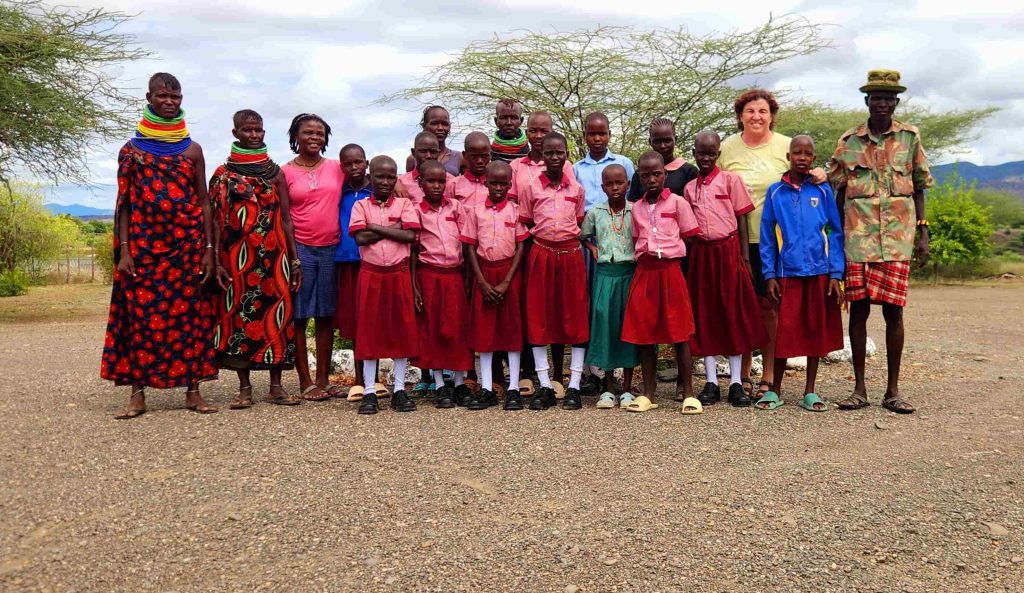 Group of schoolchildren in pink uniforms with red skirts, flanked by two adults, outdoors on a gravel lot with trees and hills in the background.